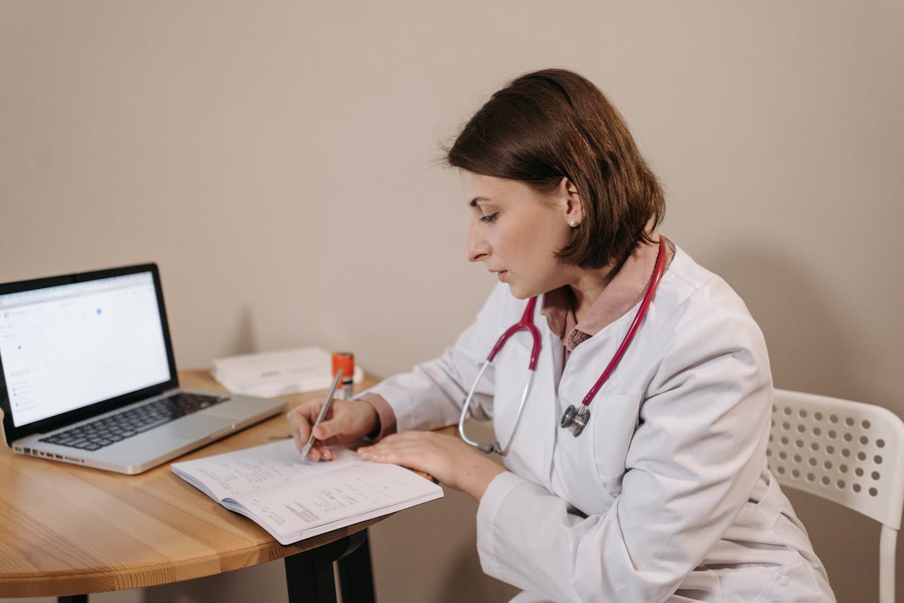 our-story-01 A female doctor in a lab coat writing a prescription in a clinic office setting with a laptop on the table.